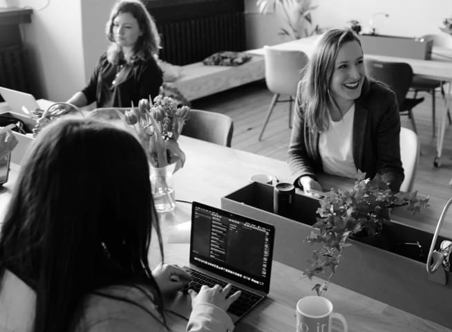 Three white women in a table. One of them is laughing and the other two staring at a laptop screen.